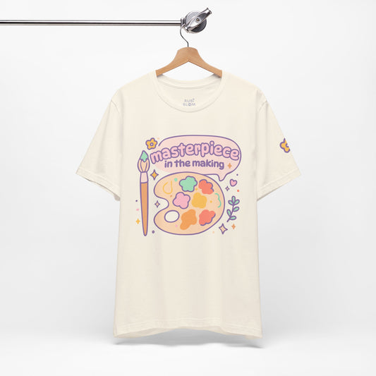 Cream “Masterpiece in the Making” pastel t-shirt hanging on rack — cozy artistic tee with colorful paint palette, cute aesthetic gift for dreamers and artists.