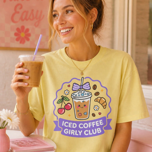 A woman sitting in a cafe setting wearing a pale yellow graphic t-shirt that reads 'ICED COFFEE GIRLY CLUB' with a cartoon iced coffee and cherries, holding an iced coffee.