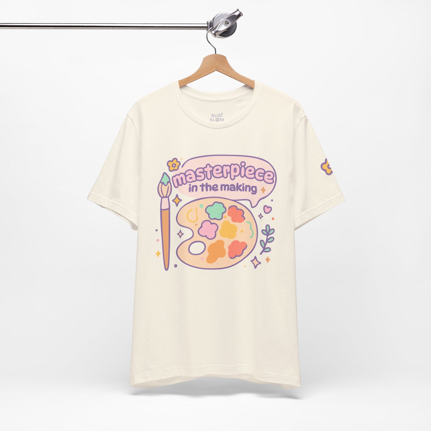 Cream “Masterpiece in the Making” pastel t-shirt hanging on rack — cozy artistic tee with colorful paint palette, cute aesthetic gift for dreamers and artists.