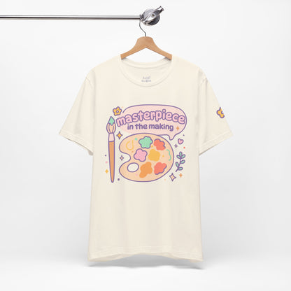 Cream “Masterpiece in the Making” pastel t-shirt hanging on rack — cozy artistic tee with colorful paint palette, cute aesthetic gift for dreamers and artists.
