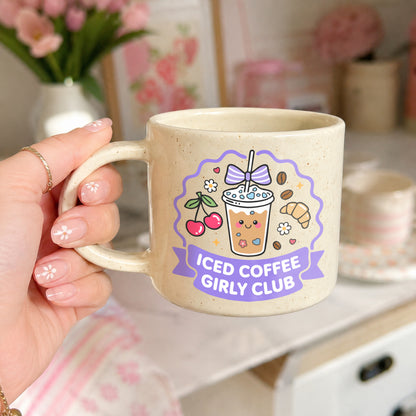 A hand holds a speckled beige ceramic mug with a cute boba coffee illustration and the text 'ICED COFFEE GIRLY CLUB' in purple. The background is a soft-focus pink and white room setting.