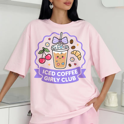 A woman wearing a light pink oversized t-shirt with a colorful cartoon graphic of an iced coffee cup, cherries, a croissant, and the text 'ICED COFFEE GIRLY CLUB' on a purple banner.