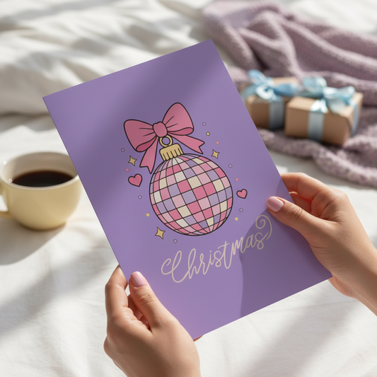 A person holds a purple Christmas greeting card featuring a pink and white disco ball ornament with a pink bow and the word 'Christmas' in white script. The card is photographed on a white bed with a cup of coffee, a blanket, and wrapped gift boxes in the background.
