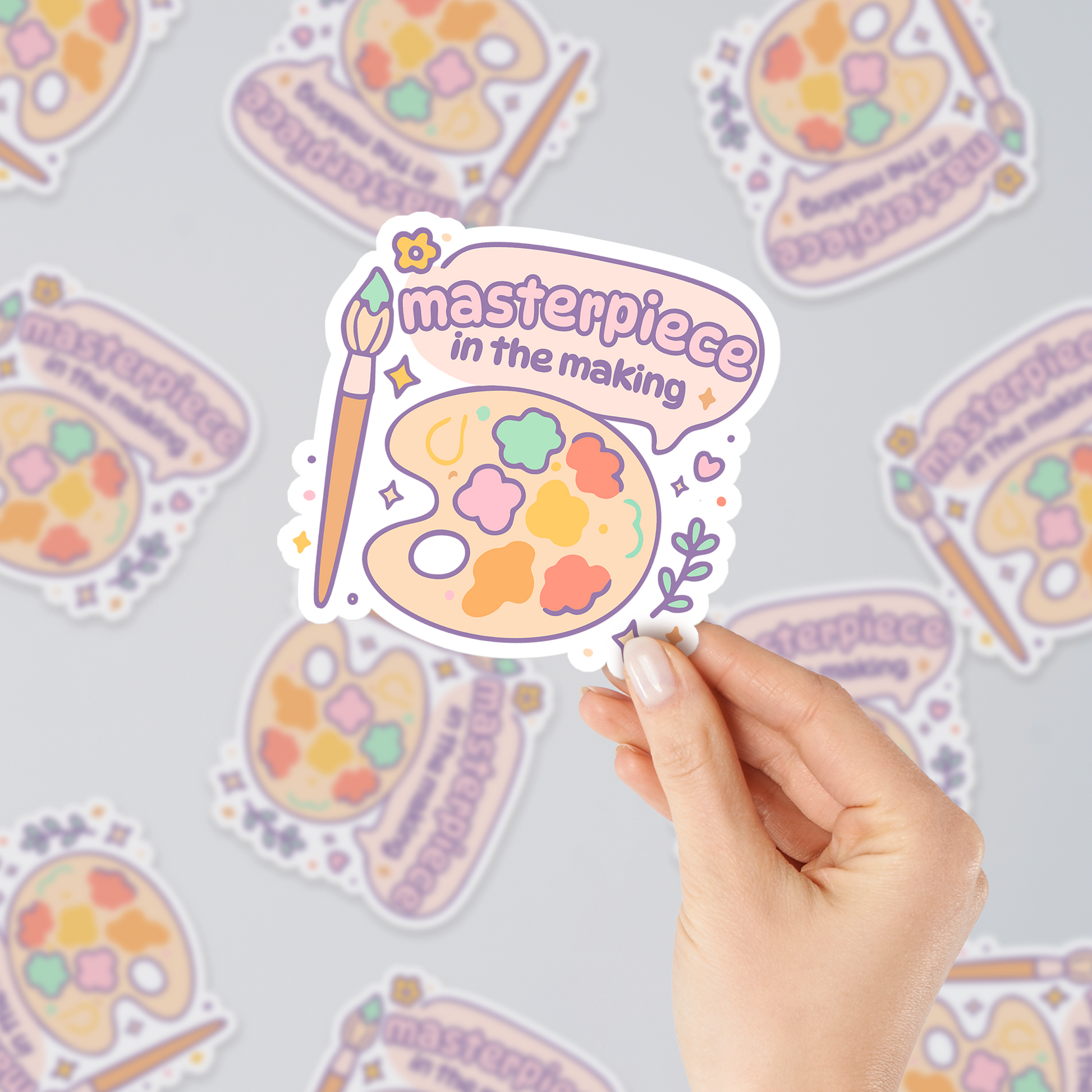 Masterpiece in the Making pastel vinyl sticker with artist palette and brush, cute aesthetic design for creatives, shown in hand with multiple stickers background.