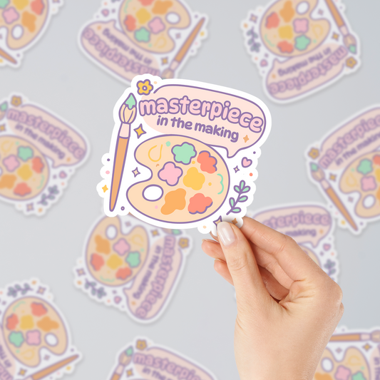 Masterpiece in the Making pastel vinyl sticker with artist palette and brush, cute aesthetic design for creatives, shown in hand with multiple stickers background.