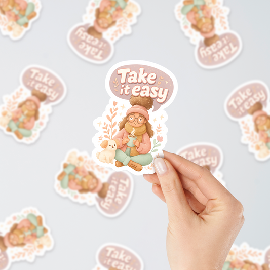 Hand holding “Take It Easy” autumn vinyl sticker with cozy girl and dog design, surrounded by multiple stickers on a light background.