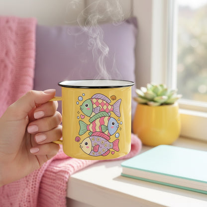 A hand holds a yellow enamel mug with a colorful fish pattern, with steam rising from the top. The mug is shown in a cozy indoor setting with a pink blanket, a purple pillow, a small potted succulent, and a notebook on a windowsill.