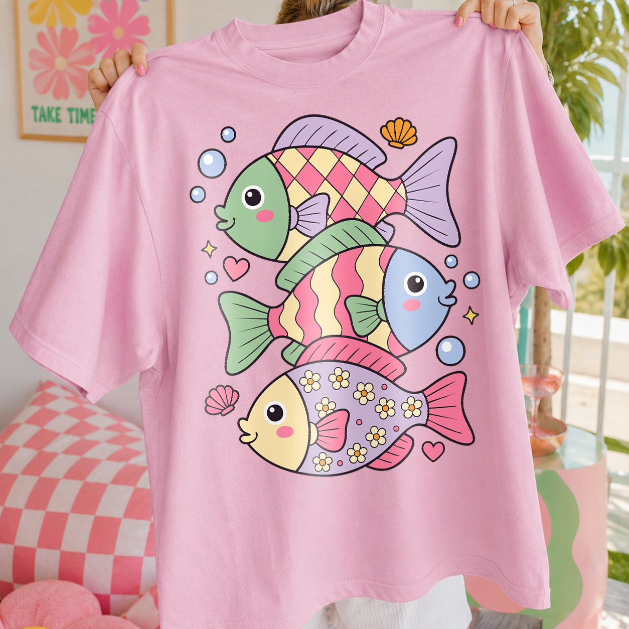 A person holds up a light pink short-sleeve T-shirt featuring a colorful cartoon graphic of three fish with pastel green, pink, blue, and yellow details, set against a softly blurred indoor background.